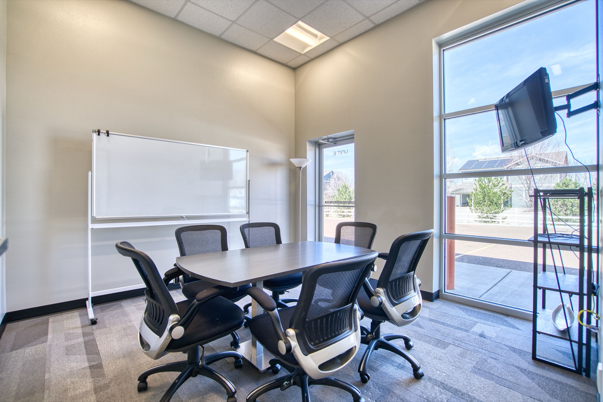 Conference room with six-seat table, rolling chairs, large whiteboard, flat-screen monitor, and floor-to-ceiling window